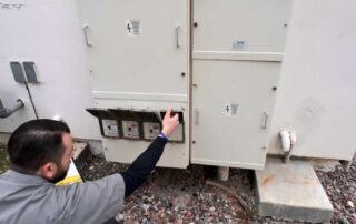 Tech in safety gear inspects outdoor electrical panels, reaching to adjust a switch.