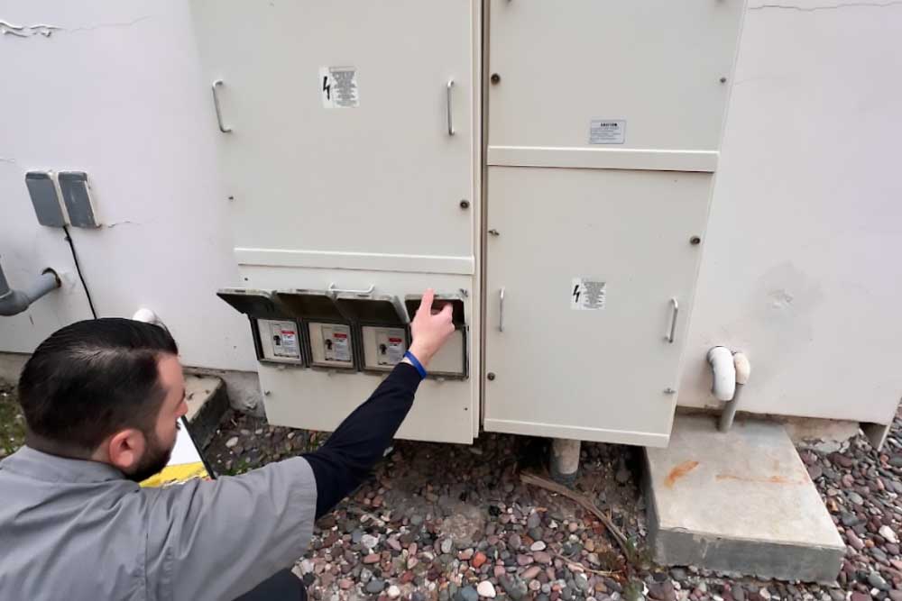 Tech in safety gear inspects outdoor electrical panels, reaching to adjust a switch.