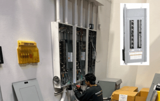 Technician working inside an open wall-mounted electrical panel; wiring and breakers visible, yellow safety device on the wall nearby.
