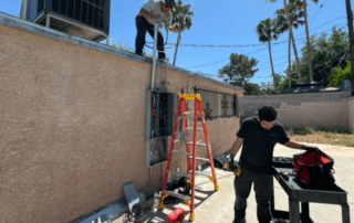 Two workers outdoors by a building; one on a ladder handling exterior electrical work, toolbox and materials on the ground.