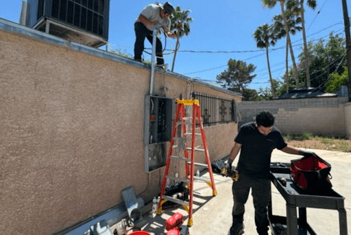 Two workers outdoors by a building; one on a ladder handling exterior electrical work, toolbox and materials on the ground.
