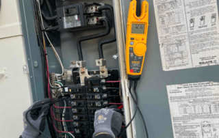 Close-up of a circuit-breaker panel with wiring; a yellow handheld testing meter rests nearby.