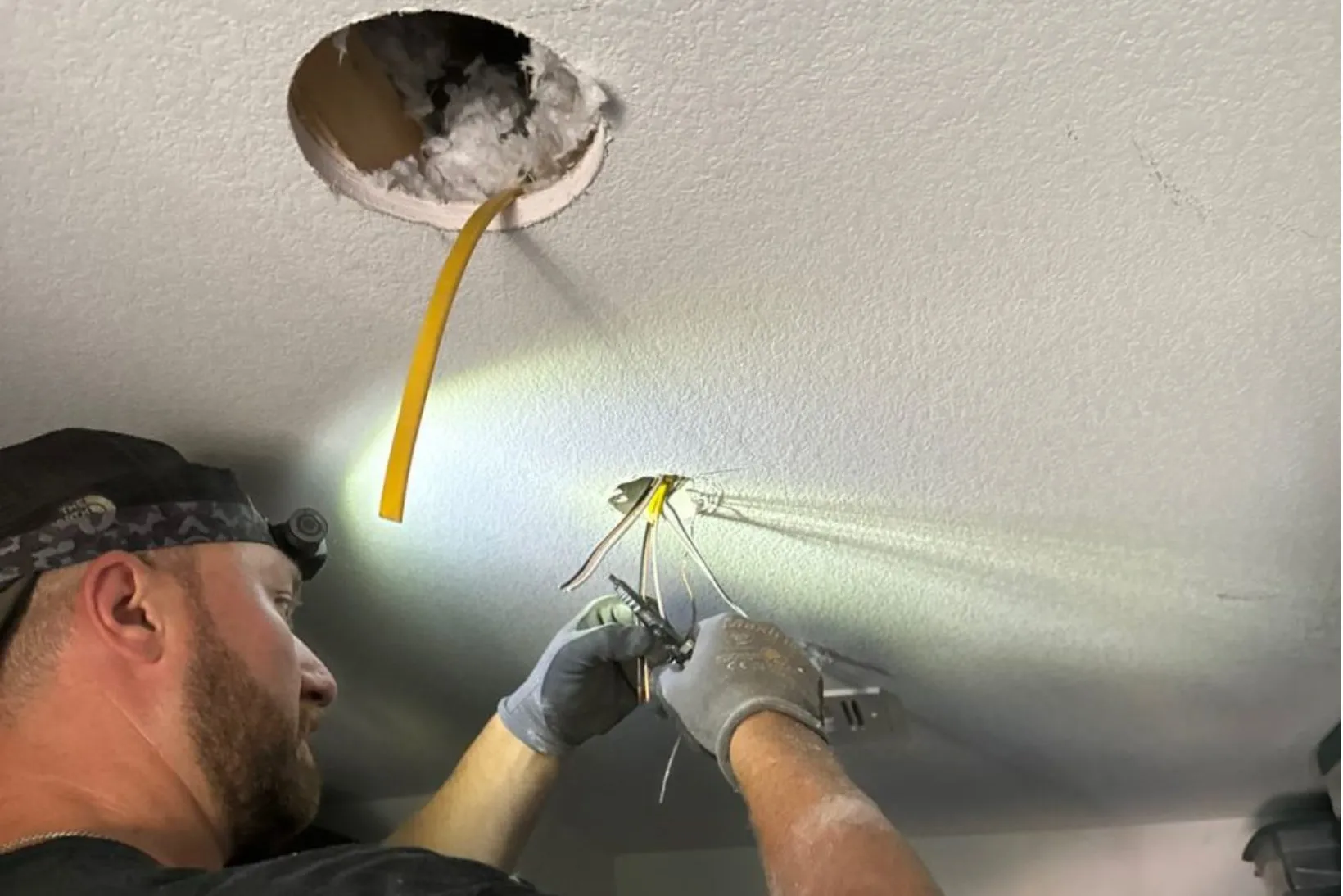 Rewiring Electrician working on electrical rewiring, connecting wires in ceiling, with yellow cable and tools visible, highlighting safety and efficiency in electrical services in Henderson.