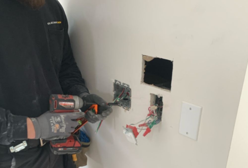 image_3 Worker wearing gloves drills into drywall with holes and exposed wiring near a wall outlet.